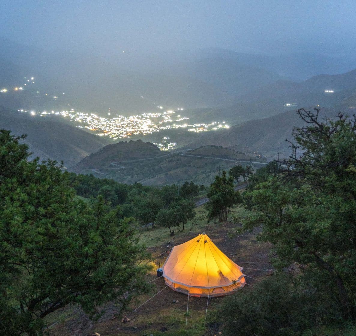 A lit tent on a hillside above a valley town at dusk